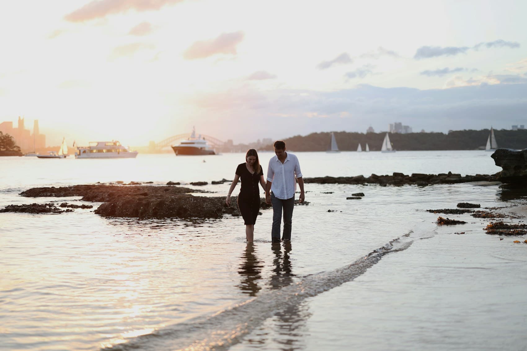 Couple walking through shallow water during sunset with boats in the distance.