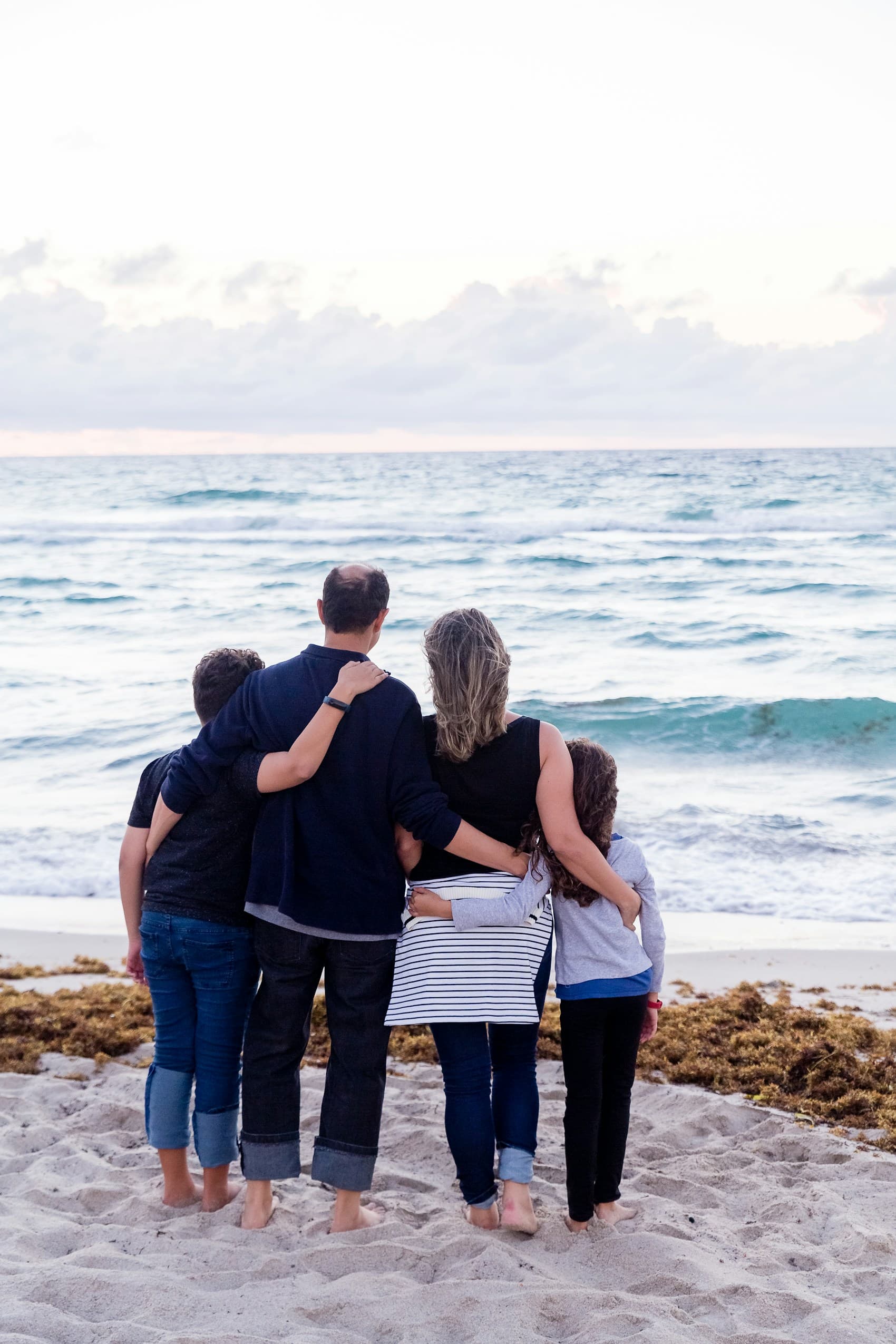 Family of four standing together on a sandy beach facing the ocean.