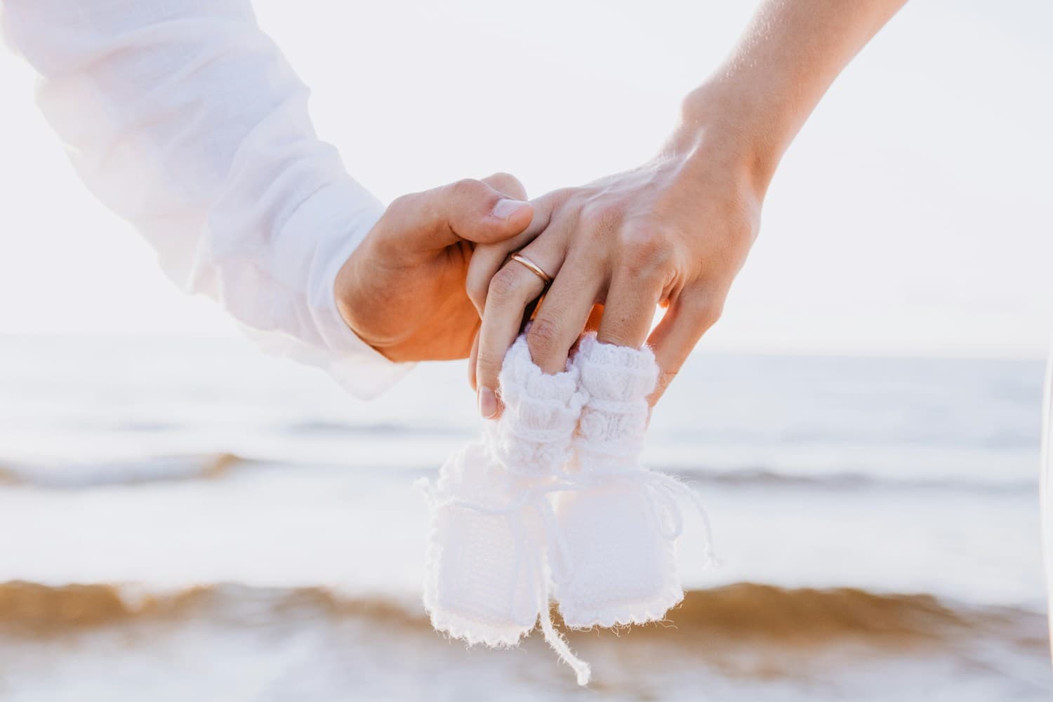 Couple holding baby shoes near the ocean with rings visible.
